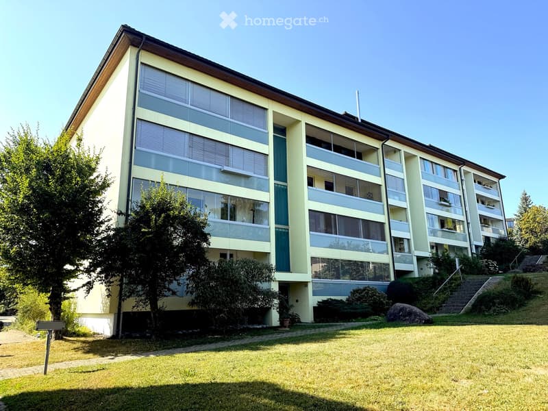 Multi-story residential building with large windows, balconies, and surrounding greenery.