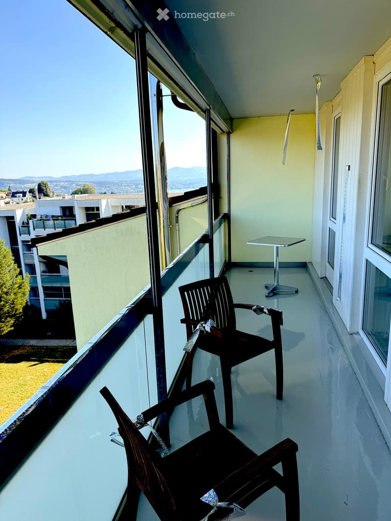 Balcony with a white floor, two wooden chairs, and a small round table, overlooking a scenic view of mountains and buildings.