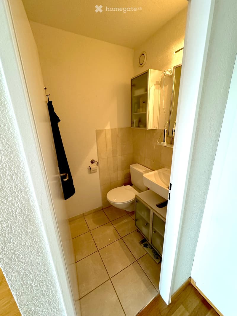 Compact bathroom with a toilet, sink, mirror, and cabinet. Beige tiled floor and walls, with a towel hanging on the door.