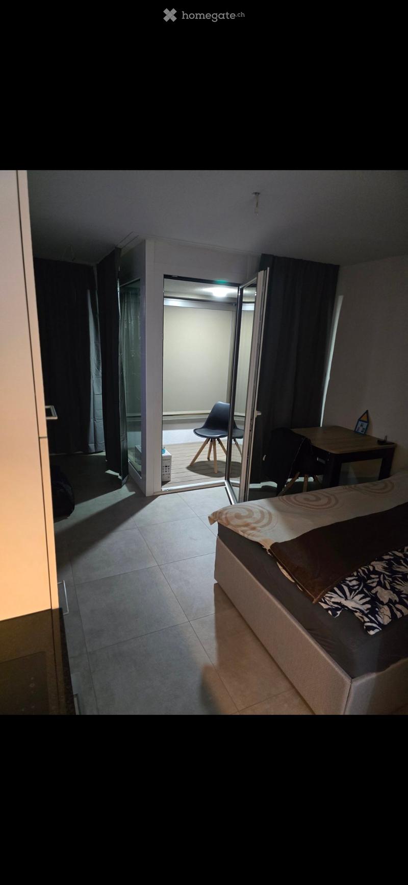 A small, modern bedroom with a bed, a chair, a small table, and a curtained window. The room has tiled flooring and an open door leading to a balcony area.