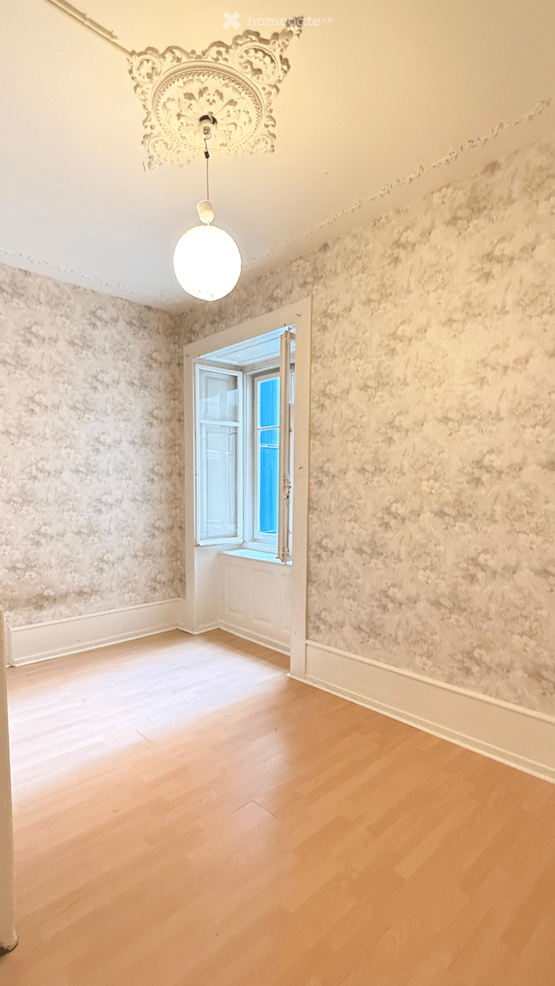 Empty room with light wooden flooring, textured walls, and a decorative ceiling light fixture. An open doorway leads to a windowed area.