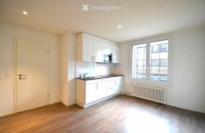 Empty kitchen with white cabinets, a microwave, sink, and countertop. Wooden flooring and large windows.