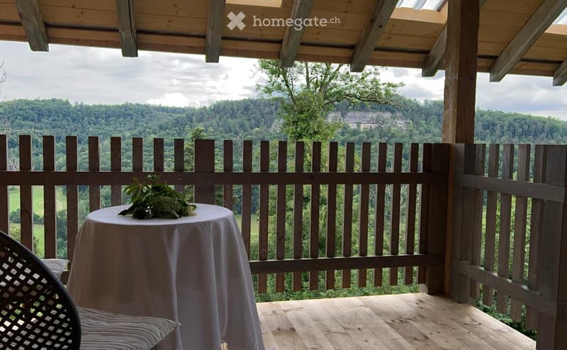 Wooden balcony with a scenic mountain view, featuring a small round table with a floral centerpiece and a wicker chair.