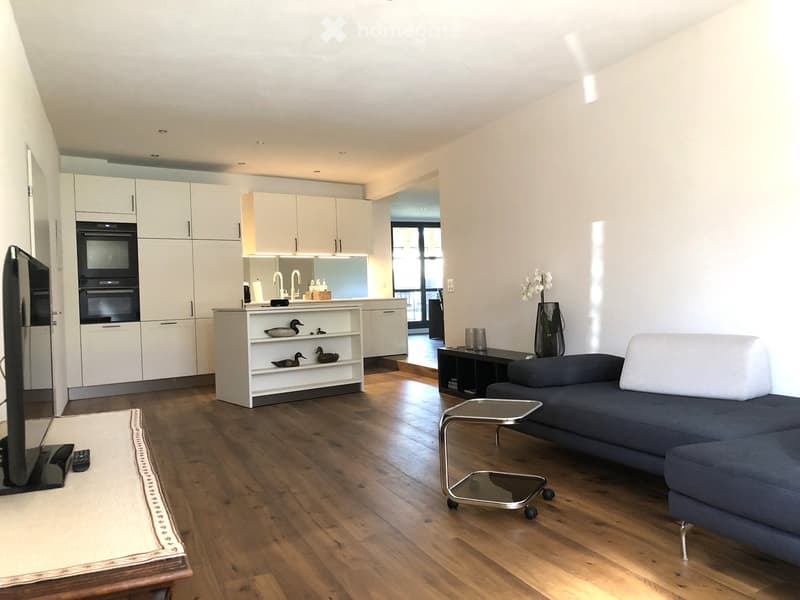 Open-plan living and dining area with a grey sofa, coffee table, TV stand, and kitchen area featuring white cabinets and an oven. Wooden flooring and modern decor.