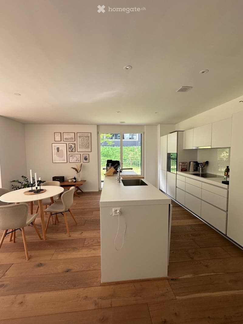 Modern open plan living area with a white kitchen island, a dining table, and a hallway leading to a garden.