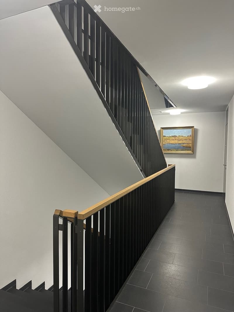 Modern staircase with black metal railings and wooden steps, tiled hallway floor, framed landscape painting on wall, ceiling lights