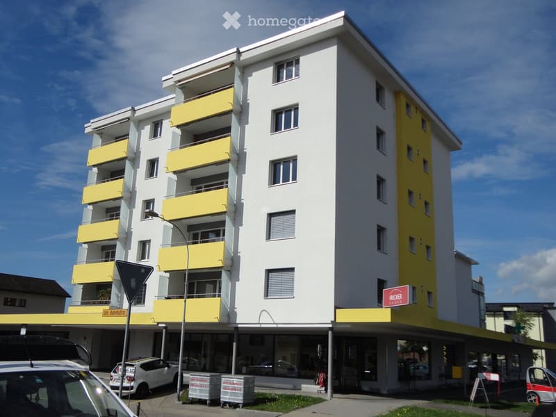 Modern multi-story residential building with white and yellow facade, multiple balconies, and ground-floor commercial spaces.
