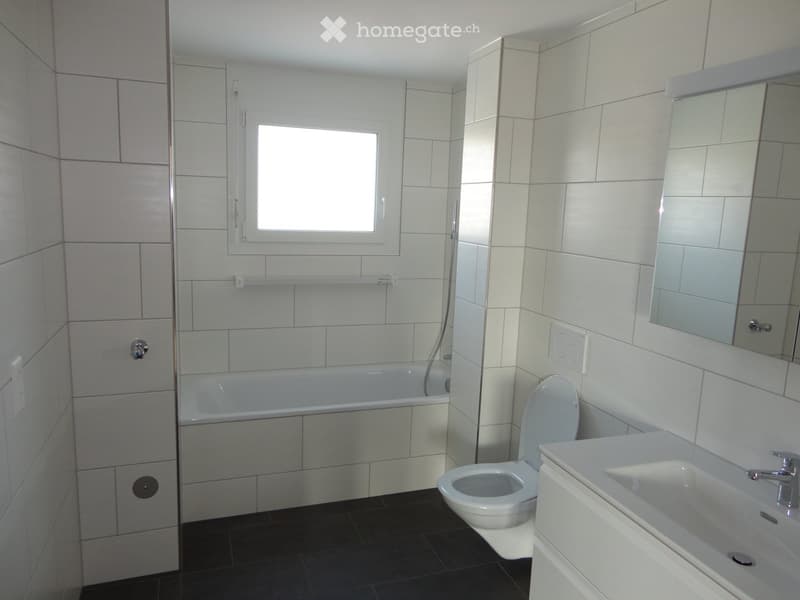 Modern bathroom with white tiles, bathtub, toilet, sink, and large mirror. Features a window for natural light and sleek fixtures.