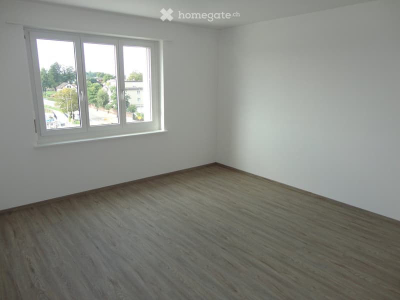 Empty room with light wooden flooring and white walls, featuring a large window with white frame and view of the street.
