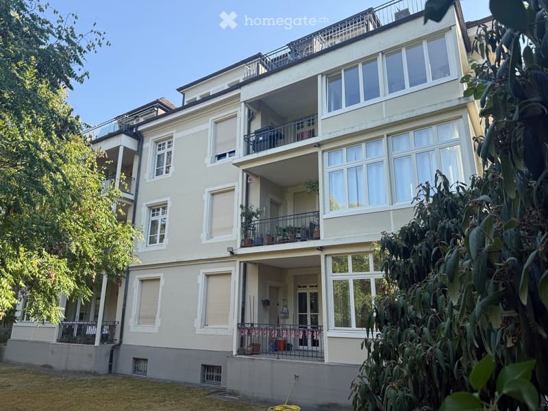 Multi-story residential building with balconies, large windows, and greenery in the foreground.