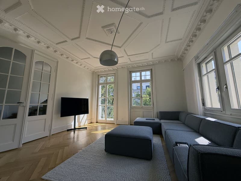 Elegant living room with ornate ceiling, large windows, grey sectional sofa, ottomans, flat-screen TV, and a modern floor lamp.