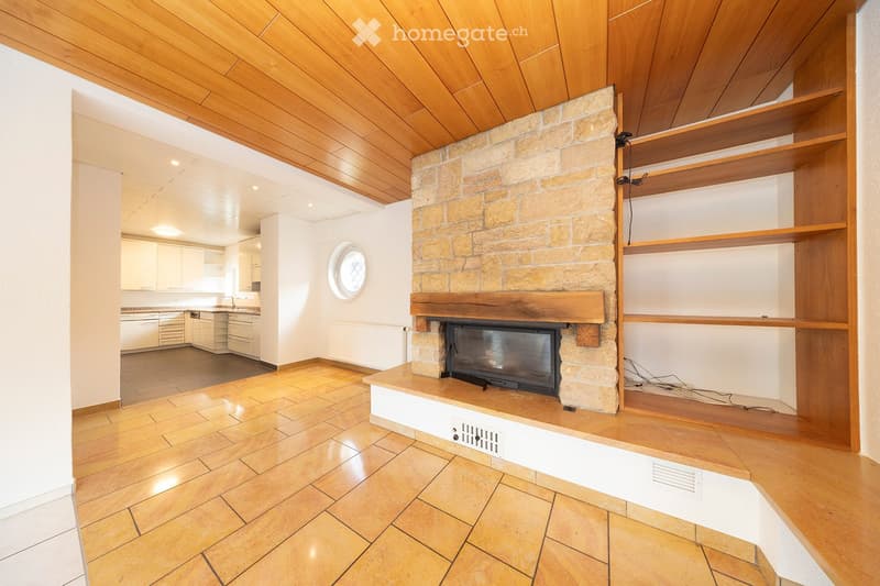 Open-plan living and dining area with a stone fireplace, wooden shelves, and a modern kitchen in the background.