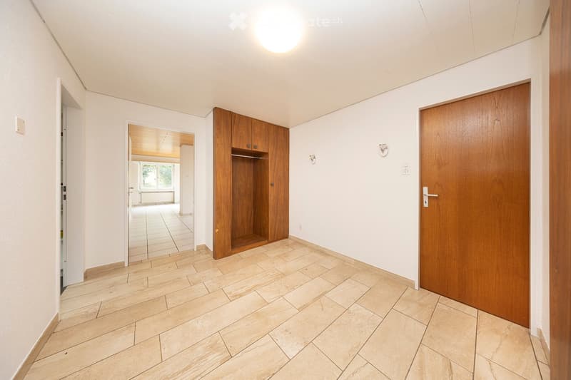 Empty hallway with light-colored tiled flooring, white walls, and wooden doors. A built-in wooden cabinet is visible on the left side, and an open doorway leads to another room with a slanted wooden ceiling and large windows.