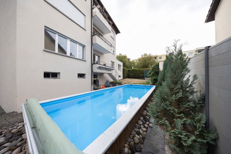 Private outdoor area with a swimming pool, surrounded by rocks and greenery, adjacent to a multi-story residential building with balconies and windows.