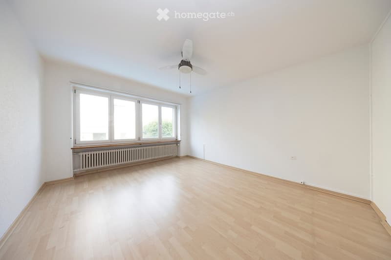 Empty room with light wooden flooring, white walls, large windows, and a ceiling fan.