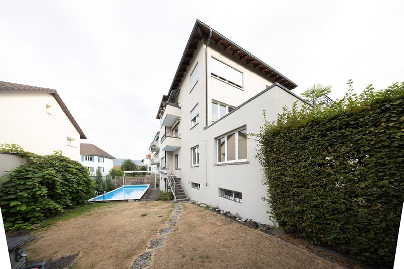 Modern multi-story building with white exterior, large windows, balconies, and a swimming pool in the garden area.