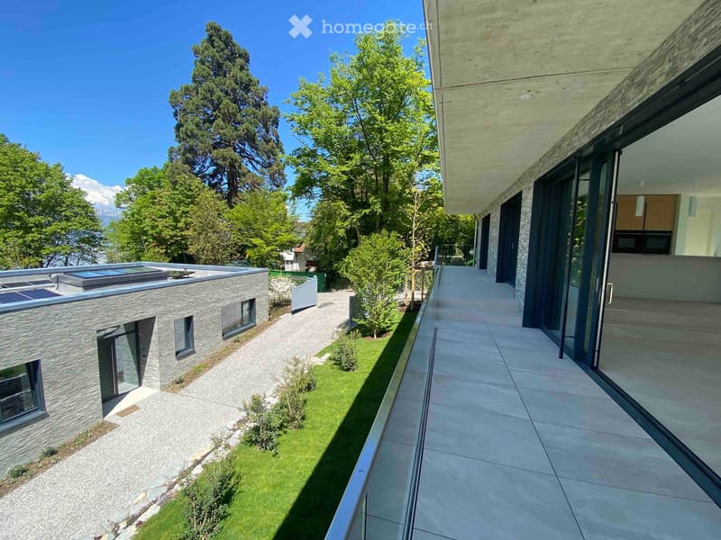 Modern house with a flat roof, large windows, and a spacious outdoor area featuring a paved walkway, grassy lawn, and surrounding trees.