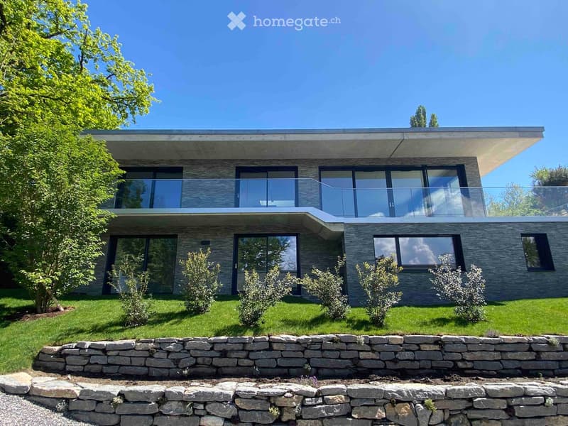 Modern two-story house with large windows, stone facade, and a raised ground floor. Features a balcony on the upper level and a stone retaining wall in the front yard.