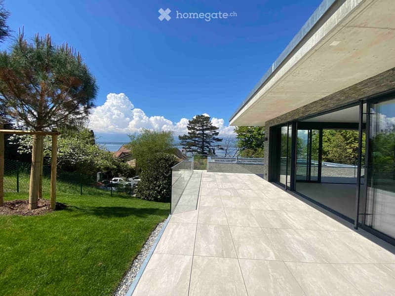 Modern outdoor patio with large glass sliding doors leading to a grassy garden, surrounded by trees and a view of the water.