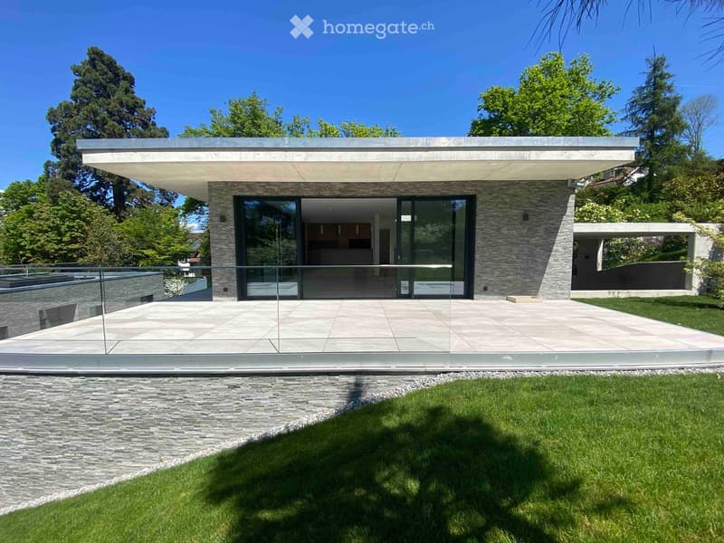 Modern two-story house with large glass windows, stone facade, and a spacious patio area surrounded by greenery.