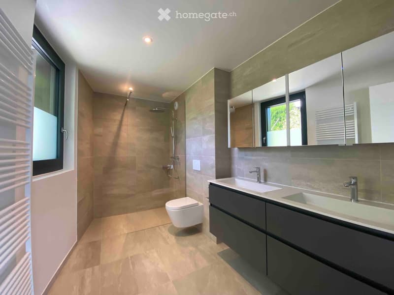 Modern bathroom with beige tiles, double sink, large mirror, toilet, and glass-enclosed shower area.
