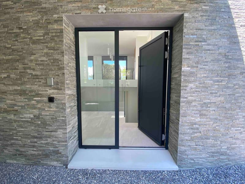 Modern entrance with a large glass door framed in black metal, flanked by textured stone walls. The door is partially open, revealing a glimpse of the interior with white walls and modern lighting.