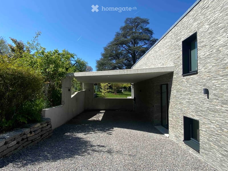 Modern stone-clad building with a flat roof and large windows, surrounded by greenery and a gravel driveway.