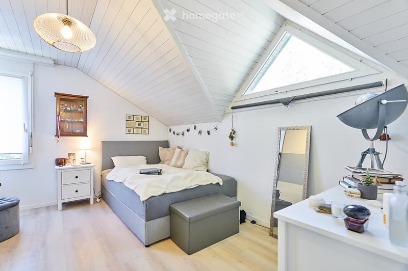 Cozy bedroom with a slanted white ceiling, wooden floor, grey bed with pillows and blanket, nightstand with lamp, large mirror, and a small table with decorative items.