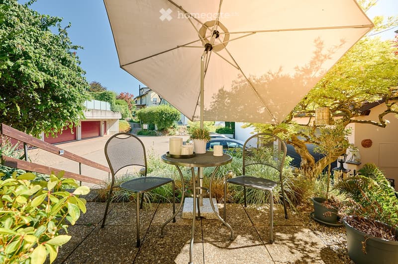 Balcony with a small round table, two metal chairs, and a large umbrella providing shade. Potted plants and a view of the street are visible.