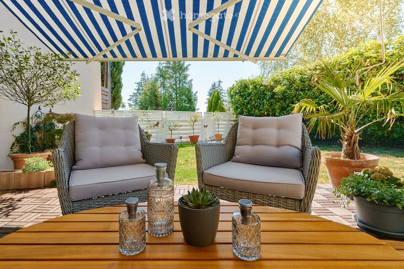 A well-maintained outdoor patio with wicker chairs, a wooden table, decorative glass bottles, and potted plants, shaded by a blue and white striped awning.