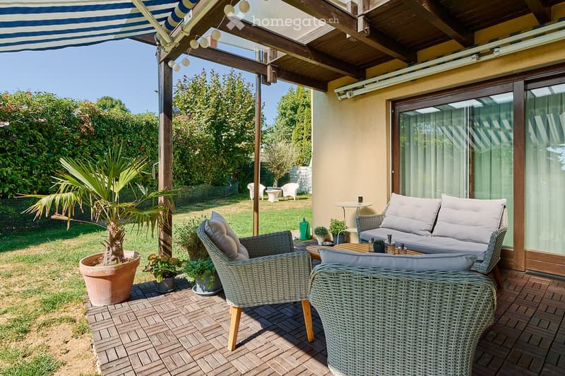 A covered outdoor patio with wicker furniture, including a sofa, chairs, and a small table, surrounded by greenery and potted plants.