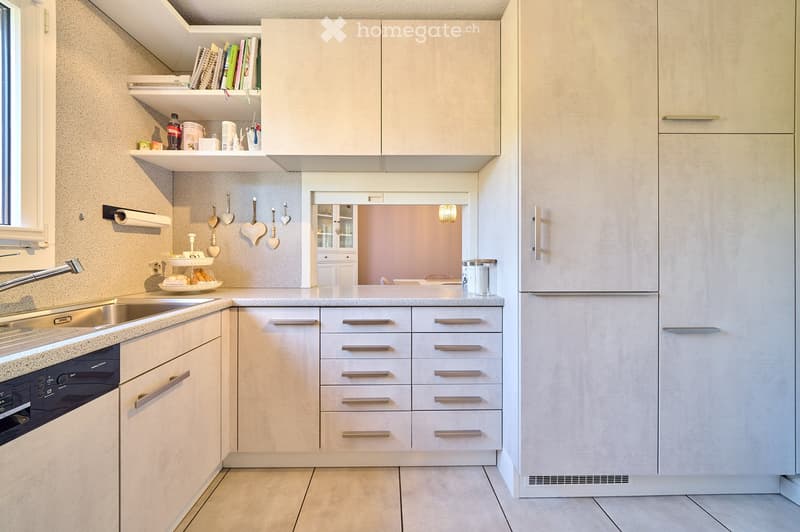 Modern kitchen with light-colored cabinets, stainless steel sink, dishwasher, and refrigerator. Shelves hold books, bottles, and small items. Decorative hearts hang on the wall, and a small window provides natural light.