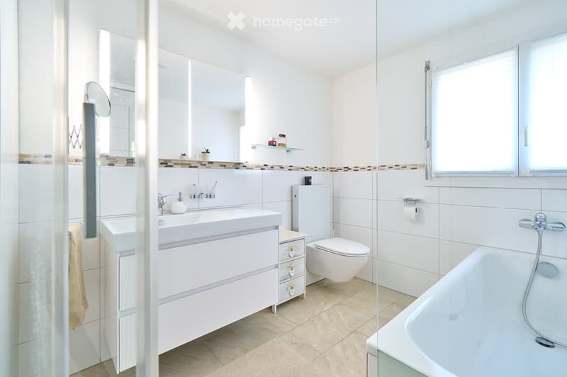 Modern bathroom with white tiles, a large sink with a cabinet, a toilet, a bathtub, and a mirror. The space is bright with natural light from a window and features a glass shower enclosure.