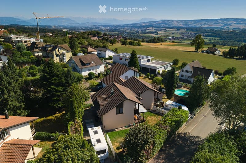 Aerial view of a suburban neighborhood with multiple houses, gardens, a swimming pool, and a scenic mountain backdrop.