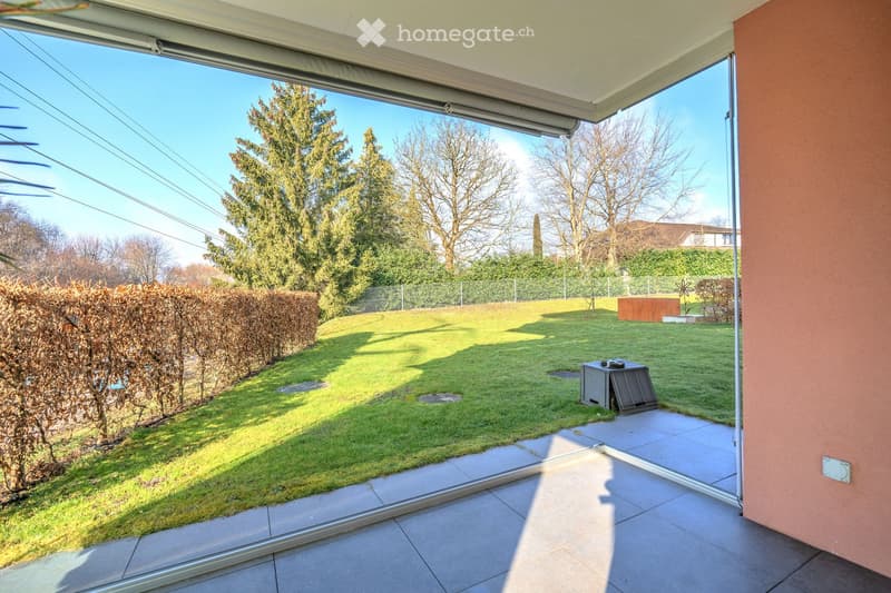 Large patio with glass walls, green lawn, hedges, small bench, and a trash can.