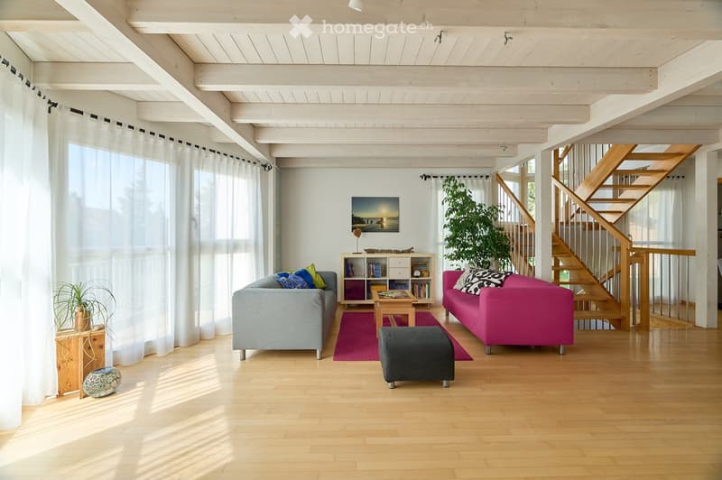 Bright living room with wooden floors, high ceilings, large windows with white curtains, two sofas, coffee table, ottoman, bookshelf, and decorative plants.