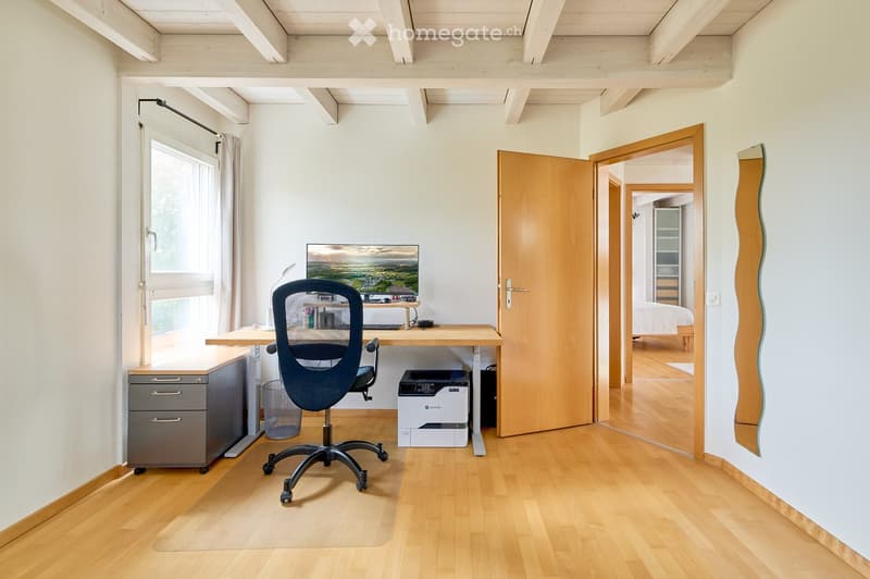 A modern, well-lit room with a wooden floor, a large window with curtains, a desk with a chair, a printer, and a screen. An open door leads to another room with a bed visible.