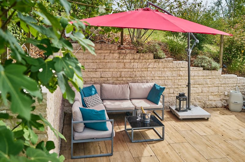 A wooden deck patio with a grey sectional sofa, teal cushions, a black coffee table, umbrella, lantern, and wine bucket.