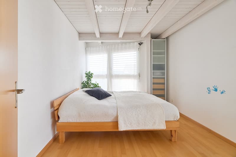A small, cozy bedroom with a wooden bed, a pillow, a wardrobe, and a small plant near the window. The room features white walls, wooden flooring, and a slanted white ceiling.