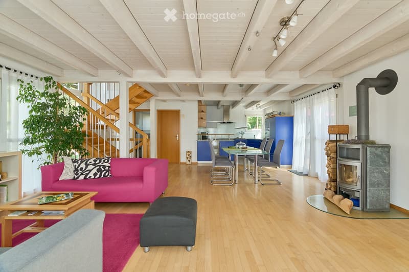 Bright living room with wooden floors, exposed wooden beams, a pink sofa, coffee table, ottoman, fireplace, dining area with a glass table and chairs, and a staircase leading to another level.