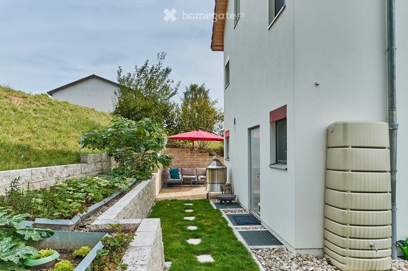 A modern garden area with a raised bed, small lawn, patio seating, and a water tank attached to the house.