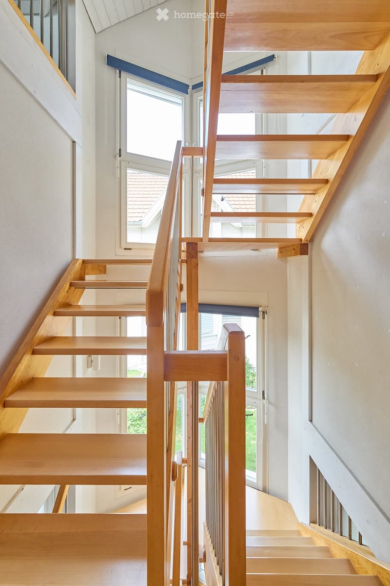 Wooden staircase with handrail and open balustrade, leading to a bright room with large windows and a view of the roof and greenery outside.