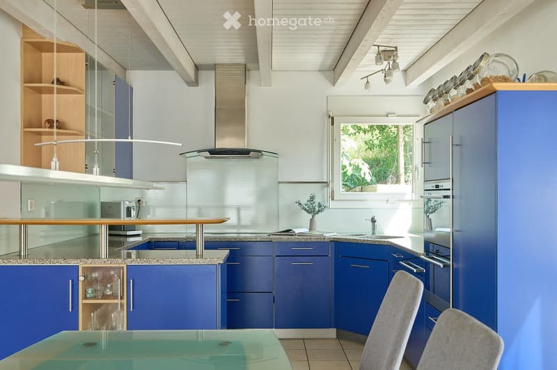 Modern kitchen with blue cabinets, granite countertops, a central island, and a dining area with a glass table and chairs. Features include a stainless steel oven, refrigerator, and a window with a view of greenery.