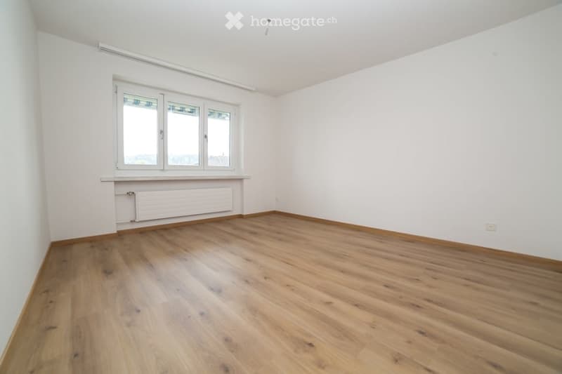 Empty room with wooden flooring, white walls, and large windows with white frames. A radiator is visible under the windows.