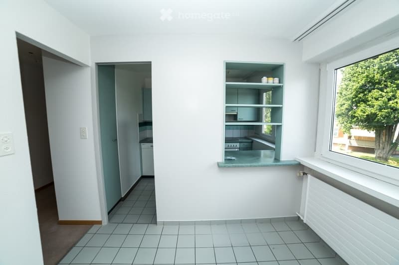 Empty room with white walls, tiled floor, and a small open kitchen area with shelves and a countertop visible through an open doorway.