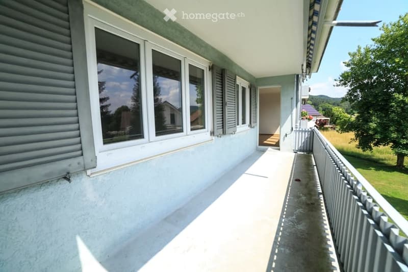 Balcony with large windows, white railing, and a view of a green landscape and mountains.