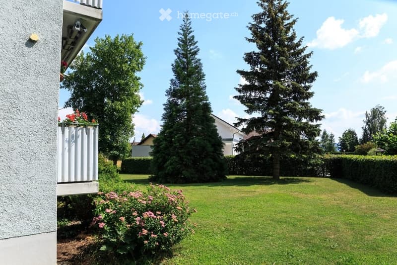 Well-maintained garden with green lawn, flower beds, tall coniferous trees, and a small balcony with flowers visible on the left side.