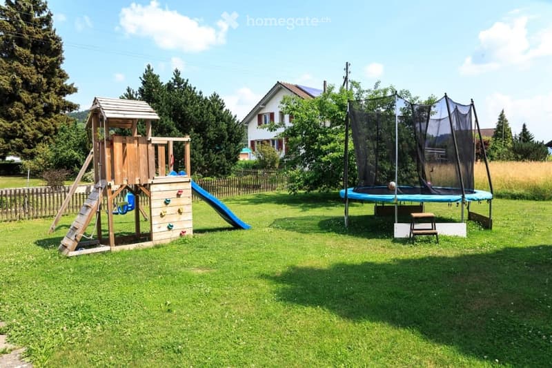 A well-maintained garden with a wooden play structure featuring a slide and climbing wall, a trampoline with a safety net, and a small bench. The area is surrounded by a wooden fence and trees, with a house visible in the background.