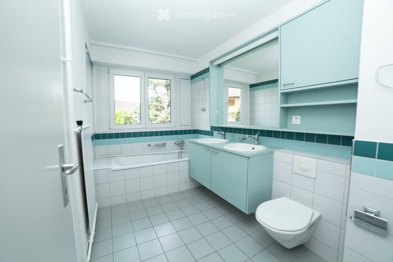 Bright bathroom with white tiles, a bathtub, double sink vanity, toilet, and a large mirror above the sink.