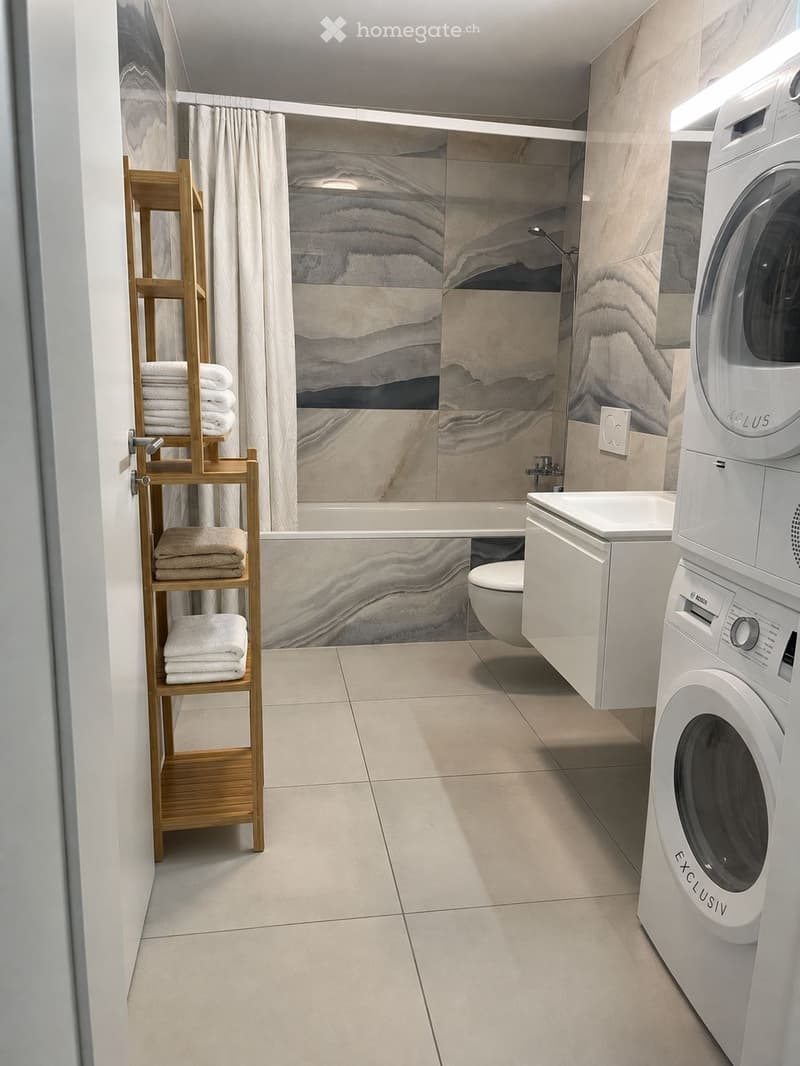 Modern bathroom with a bathtub, toilet, washing machine, and a wooden shelf holding neatly folded towels.